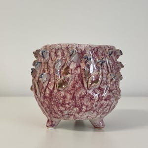 Pode incluir: Um pequeno vaso redondo de cerâmica com uma superfície texturizada roxo-avermelhada. O vaso apresenta uma base de três pés e é adornado com desenhos em relevo em forma de folha em tons de castanho, azul e branco. Ideal para plantas pequenas.