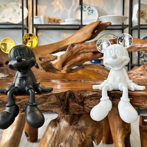 Puede incluir: Dos figuras de Mickey Mouse, una negra con orejas doradas y otra blanca con orejas plateadas, sentadas sobre una mesa de madera. Las figuras est&aacute;n en una pose relajada, con las piernas colgando. Se ven platos decorativos en el fondo.