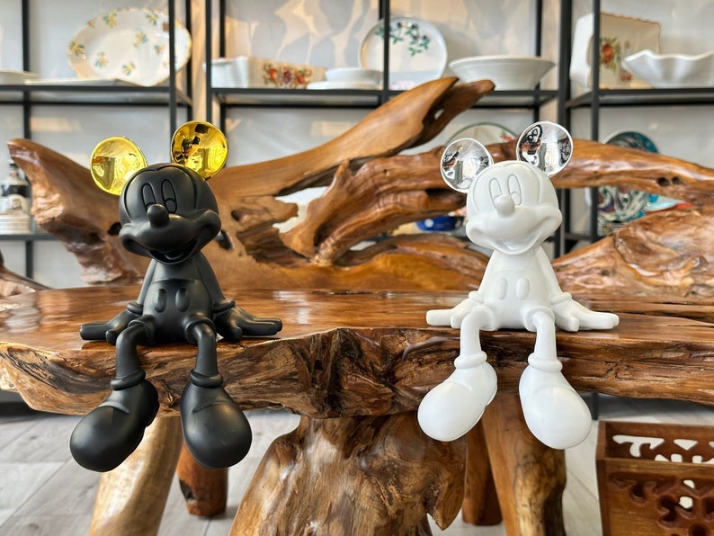 Puede incluir: Dos figuras de Mickey Mouse, una negra con orejas doradas y otra blanca con orejas plateadas, sentadas sobre una mesa de madera. Las figuras est&aacute;n en una pose relajada, con las piernas colgando. Se ven platos decorativos en el fondo.
