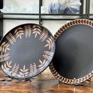 Könnte beinhalten: Zwei schwarze Keramikplatten mit Blattmustern. Eine Platte hat einen Blattrand, die andere Blätter am Rand. Die Teller werden auf Metallständern präsentiert, die ihre dekorativen Muster und die dunkle Farbe hervorheben.