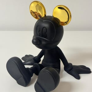 Pu&ograve; includere: Una statuetta di Topolino nera con orecchie dorate. La statuetta &egrave; seduta con le braccia e le gambe distese. La figura ha una finitura opaca ed &egrave; posta su uno sfondo bianco.