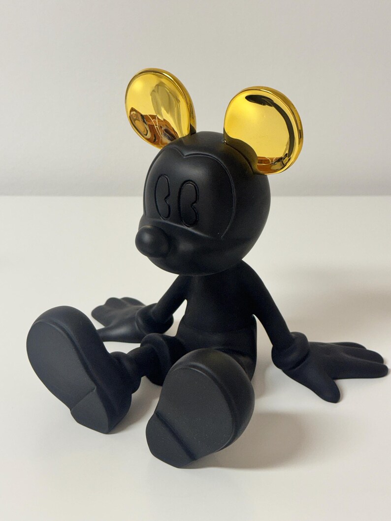 Pu&ograve; includere: Una statuetta di Topolino nera con orecchie dorate. La statuetta &egrave; seduta con le braccia e le gambe distese. La figura ha una finitura opaca ed &egrave; posta su uno sfondo bianco.