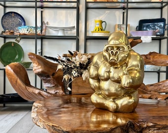 May include: A golden gorilla statue in a seated position, with a contemplative expression. The sculpture is displayed on a polished wooden surface, with shelves of decorative items in the background. The gorilla is a striking decorative piece.