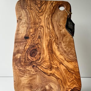 May include: A rectangular wooden cutting board with a natural wood grain pattern. The board has a handle with a hole and a tag attached. The wood grain features swirls of brown and tan colors. The board is supported by two silver metal feet.