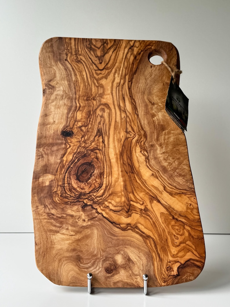 May include: A rectangular wooden cutting board with a natural wood grain pattern. The board has a handle with a hole and a tag attached. The wood grain features swirls of brown and tan colors. The board is supported by two silver metal feet.