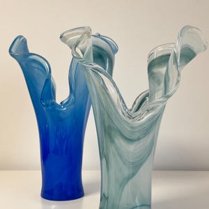 Pode incluir: Dois vasos de vidro com um design único de borda ondulada. Um é azul liso, o outro apresenta um padrão de redemoinho azul e branco. Os vasos têm uma forma alta e esguia.