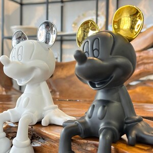 Puede incluir: Dos figuras de Mickey Mouse, una blanca con orejas plateadas y otra negra con orejas doradas, est&aacute;n sentadas una al lado de la otra. Las figuras tienen un acabado brillante y est&aacute;n posadas con las piernas extendidas. El fondo est&aacute; borroso.