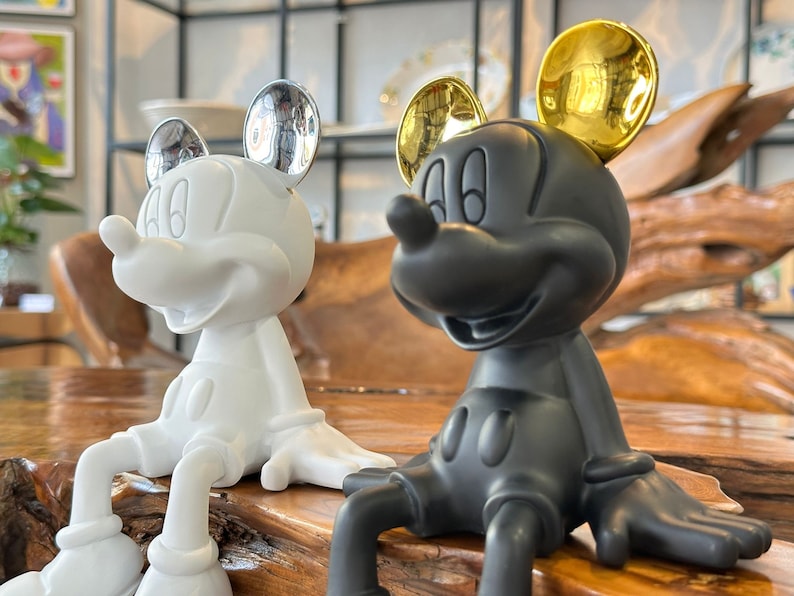 Puede incluir: Dos figuras de Mickey Mouse, una blanca con orejas plateadas y otra negra con orejas doradas, est&aacute;n sentadas una al lado de la otra. Las figuras tienen un acabado brillante y est&aacute;n posadas con las piernas extendidas. El fondo est&aacute; borroso.