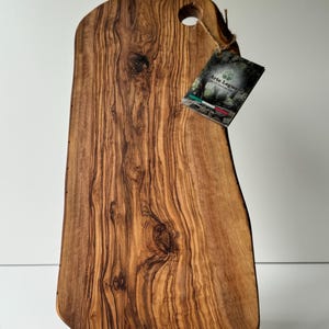 May include: A wooden cutting board with a natural edge and a hole for hanging. The board is made of olive wood and has a rich, brown color with unique grain patterns. A tag with the text "Arte Legno" is attached.