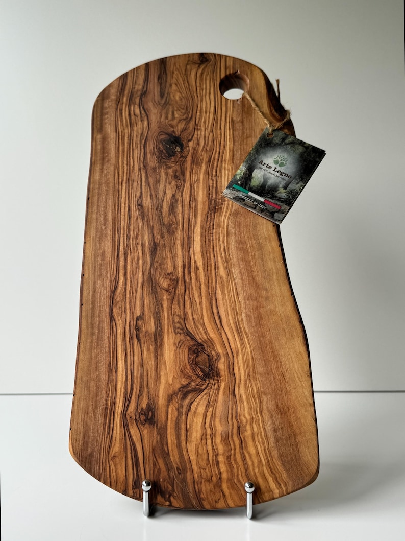 May include: A wooden cutting board with a natural edge and a hole for hanging. The board is made of olive wood and has a rich, brown color with unique grain patterns. A tag with the text "Arte Legno" is attached.