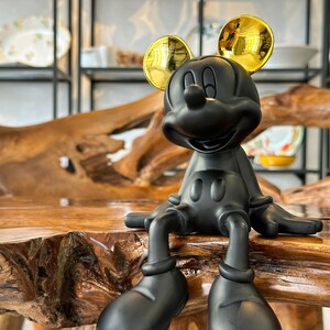 Puede incluir: Una figura negra de Mickey Mouse con orejas doradas sentada sobre una superficie de madera. La figura tiene un dise&ntilde;o cl&aacute;sico, con zapatos negros grandes y una cara sonriente. El fondo incluye estantes con platos decorativos.
