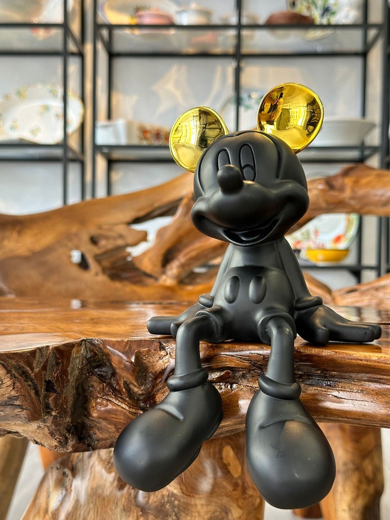 Puede incluir: Una figura negra de Mickey Mouse con orejas doradas sentada sobre una superficie de madera. La figura tiene un dise&ntilde;o cl&aacute;sico, con zapatos negros grandes y una cara sonriente. El fondo incluye estantes con platos decorativos.