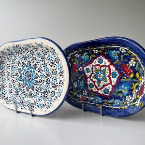 Könnte beinhalten: Zwei ovale Keramik-Servierplatten mit aufwendigen Blumenmustern. Eine Platte hat ein blau-weißes Muster, die andere ein lebhaftes Blumenmotiv mit roten, blauen und gelben Akzenten. Beide werden auf Metallständern präsentiert.