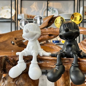 Puede incluir: Dos figuras de Mickey Mouse, una blanca y otra negra, se muestran juntas. La figura blanca tiene orejas plateadas, mientras que la negra tiene orejas doradas. Ambas est&aacute;n sentadas con las piernas extendidas, sobre una superficie de madera.