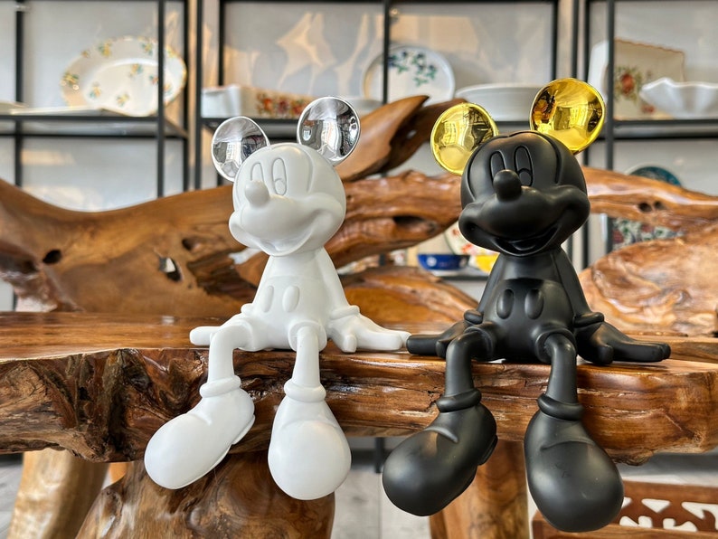 Puede incluir: Dos figuras de Mickey Mouse, una blanca y otra negra, se muestran juntas. La figura blanca tiene orejas plateadas, mientras que la negra tiene orejas doradas. Ambas est&aacute;n sentadas con las piernas extendidas, sobre una superficie de madera.