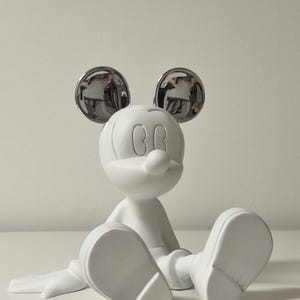 Pu&ograve; includere: Una statuetta bianca di Topolino seduto con le gambe distese. Le orecchie sono cromate, riflettendo l'ambiente circostante. La statuetta ha una finitura liscia e opaca ed &egrave; un personaggio classico Disney.