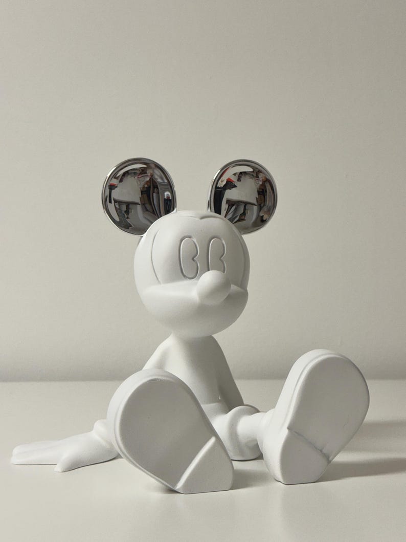 Pu&ograve; includere: Una statuetta bianca di Topolino seduto con le gambe distese. Le orecchie sono cromate, riflettendo l'ambiente circostante. La statuetta ha una finitura liscia e opaca ed &egrave; un personaggio classico Disney.