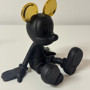 Pu&ograve; includere: Una statuetta nera di un topo dei cartoni animati con orecchie color oro, seduto con le gambe distese. Il topo ha una testa rotonda, grandi orecchie e un piccolo naso. La statuetta &egrave; su una superficie bianca.