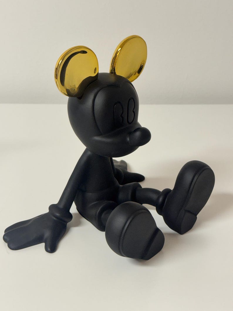 Pu&ograve; includere: Una statuetta nera di un topo dei cartoni animati con orecchie color oro, seduto con le gambe distese. Il topo ha una testa rotonda, grandi orecchie e un piccolo naso. La statuetta &egrave; su una superficie bianca.