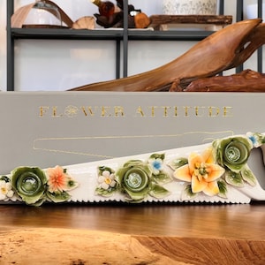 Pode incluir: Uma serra de cerâmica branca decorada com detalhes florais coloridos. A lâmina da serra apresenta flores verdes, laranja e brancas com detalhes azuis. A pega é branca. A caixa atrás da serra diz "FLOWER ATTITUDE".