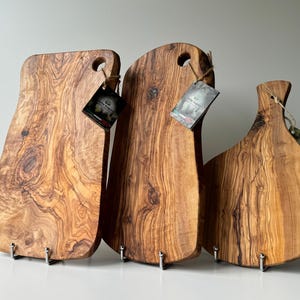 May include: Three wooden cutting boards with natural wood grain patterns. Each board has a unique shape and a hole for hanging. The boards are displayed on metal stands, showcasing their rustic charm.