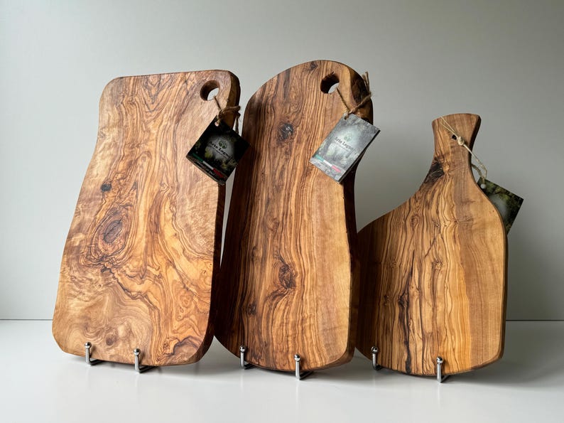 May include: Three wooden cutting boards with natural wood grain patterns. Each board has a unique shape and a hole for hanging. The boards are displayed on metal stands, showcasing their rustic charm.