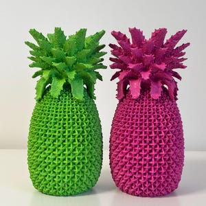 Op de afbeelding: Twee decoratieve ananas sculpturen. Eén is felgroen, de andere is fel magenta. Beide hebben een textuur en een stekelige kroon. De ananassen staan naast elkaar tegen een witte achtergrond.