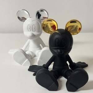 Pu&ograve; includere: Due statuette di Topolino. Una &egrave; bianca con orecchie argentate, l'altra &egrave; nera con orecchie dorate. Entrambe sono in posizione seduta con le braccia e le gambe distese. Le statuette sono su una superficie bianca.