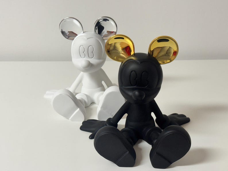 Pu&ograve; includere: Due statuette di Topolino. Una &egrave; bianca con orecchie argentate, l'altra &egrave; nera con orecchie dorate. Entrambe sono in posizione seduta con le braccia e le gambe distese. Le statuette sono su una superficie bianca.