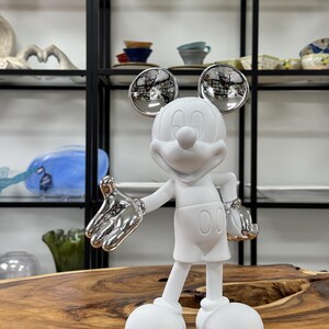 Może przedstawiać: Biała ceramiczna figurka Myszki Miki z srebrnymi akcentami. Figurka stoi z wyciągniętymi rękami, a głowa jest lekko przechylona na bok. Figurka ma około 30 cm wysokości.