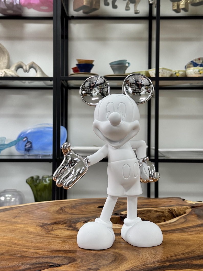 Może przedstawiać: Biała ceramiczna figurka Myszki Miki z srebrnymi akcentami. Figurka stoi z wyciągniętymi rękami, a głowa jest lekko przechylona na bok. Figurka ma około 30 cm wysokości.