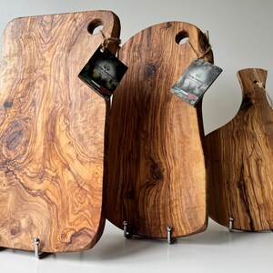 May include: Three wooden cutting boards with natural wood grain patterns. Each board has a unique shape and a hole for hanging. The boards are various shades of brown and are displayed on metal stands. Tags are attached to each board.