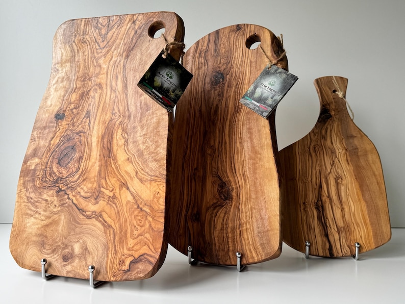 May include: Three wooden cutting boards with natural wood grain patterns. Each board has a unique shape and a hole for hanging. The boards are various shades of brown and are displayed on metal stands. Tags are attached to each board.