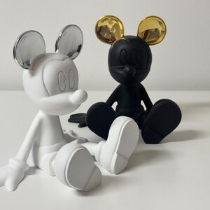 Pu&ograve; includere: Due statuette stilizzate di Topolino sedute. Una &egrave; bianca con orecchie argentate, l'altra nera con orecchie dorate. Sono su una superficie bianca. Design moderno.
