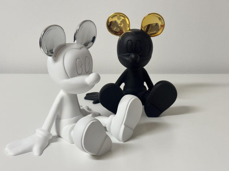 Pu&ograve; includere: Due statuette stilizzate di Topolino sedute. Una &egrave; bianca con orecchie argentate, l'altra nera con orecchie dorate. Sono su una superficie bianca. Design moderno.