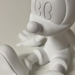 Pu&ograve; includere: Una statuetta bianca di un topo dei cartoni animati con grandi orecchie, un naso rotondo e pantaloncini. Il topo &egrave; seduto con le gambe distese. La statuetta &egrave; realizzata in un materiale liscio e opaco.