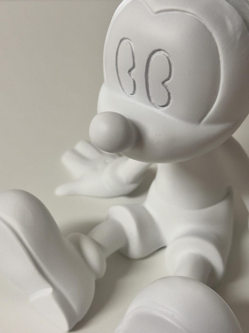Pu&ograve; includere: Una statuetta bianca di un topo dei cartoni animati con grandi orecchie, un naso rotondo e pantaloncini. Il topo &egrave; seduto con le gambe distese. La statuetta &egrave; realizzata in un materiale liscio e opaco.
