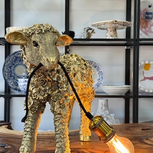 May include: A gold-coloured lamb-shaped lamp with a black cord and a lit lightbulb. The lamb is decorated with floral patterns and stands on a wooden surface. The lamp is a decorative item.