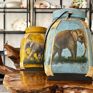 May include: Two decorative, cylindrical containers with elephant illustrations. One is yellow, the other is blue. Both have a woven border and a black base. The elephant art is brown and gray, set against a background of green grass and a light blue or yellow sky.