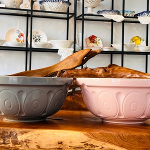 May include: Two large ceramic mixing bowls, one gray and one pink, with a decorative design of overlapping circles. The bowls are sitting on a wooden surface.