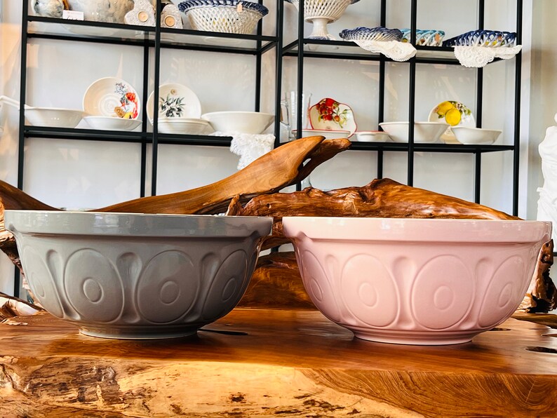 May include: Two large ceramic mixing bowls, one gray and one pink, with a decorative design of overlapping circles. The bowls are sitting on a wooden surface.