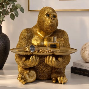 Könnte beinhalten: Eine goldfarbene Gorillafigur in sitzender Position, die ein flaches, kreisförmiges goldenes Tablett hält. Auf dem Tablett befinden sich eine goldfarbene Armbanduhr, eine goldene Kette und ein quadratischer Glasparfümflakon mit dem Etikett RITUALS.