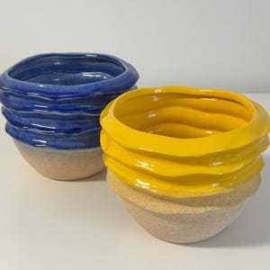 Op de afbeelding: Twee keramische kommen met golvende, geribbelde randen. De ene kom heeft een glanzend, levendig blauw glazuur aan de bovenkant, terwijl de andere een glanzend, felgeel glazuur heeft. Beide kommen hebben een gespikkelde, matte, lichtbeige basis.