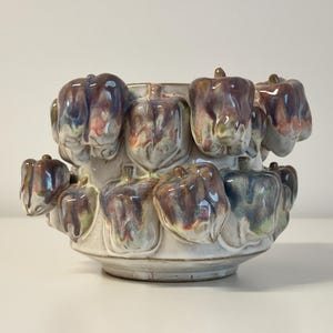 May include: A ceramic vase with a unique design featuring multiple bell pepper shapes in shades of purple, blue, and green. The vase has a cream-colored base and is suitable for decorative purposes.