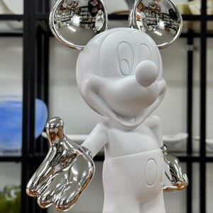 Może przedstawiać: Biała ceramiczna figurka Myszki Miki z srebrnymi metalowymi uszami i rękami. Figurka stoi z wyciągniętymi rękami.