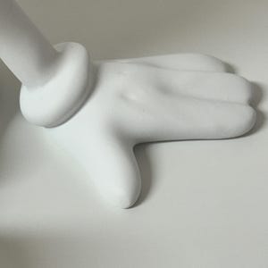 Pu&ograve; includere: Una scultura di mano bianca stilizzata con un polso arrotondato e un dettaglio ad anello. Le dita sono leggermente curve e il design generale &egrave; liscio e minimalista. La scultura &egrave; ambientata su uno sfondo bianco.