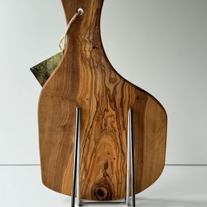 May include: A wooden cutting board with a natural wood grain pattern, displayed on a chrome stand. The board has a handle with a hole for hanging and a tag attached. The wood tones range from light to dark brown.