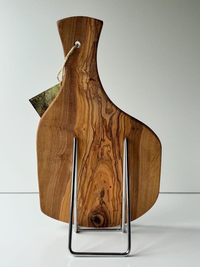 May include: A wooden cutting board with a natural wood grain pattern, displayed on a chrome stand. The board has a handle with a hole for hanging and a tag attached. The wood tones range from light to dark brown.