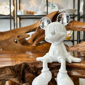 Puede incluir: Una figura blanca de Mickey Mouse con orejas reflectantes sentada sobre una superficie de madera. La figura est&aacute; en posici&oacute;n sentada, con las piernas colgando. El fondo presenta estantes con varios platos decorativos.