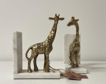 May include: Two brass giraffe figurines, one standing and one facing, sit on white marble bookends. The giraffes have detailed patterns and are connected to the bookends. The bookends are rectangular with a vertical support.